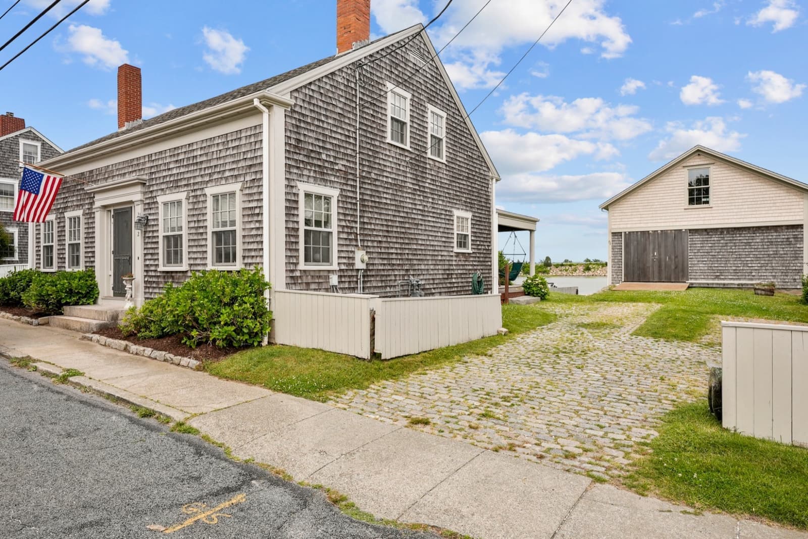 House with cobblestone driveway, water visible behind, showing the relationship between house and outbuildings
